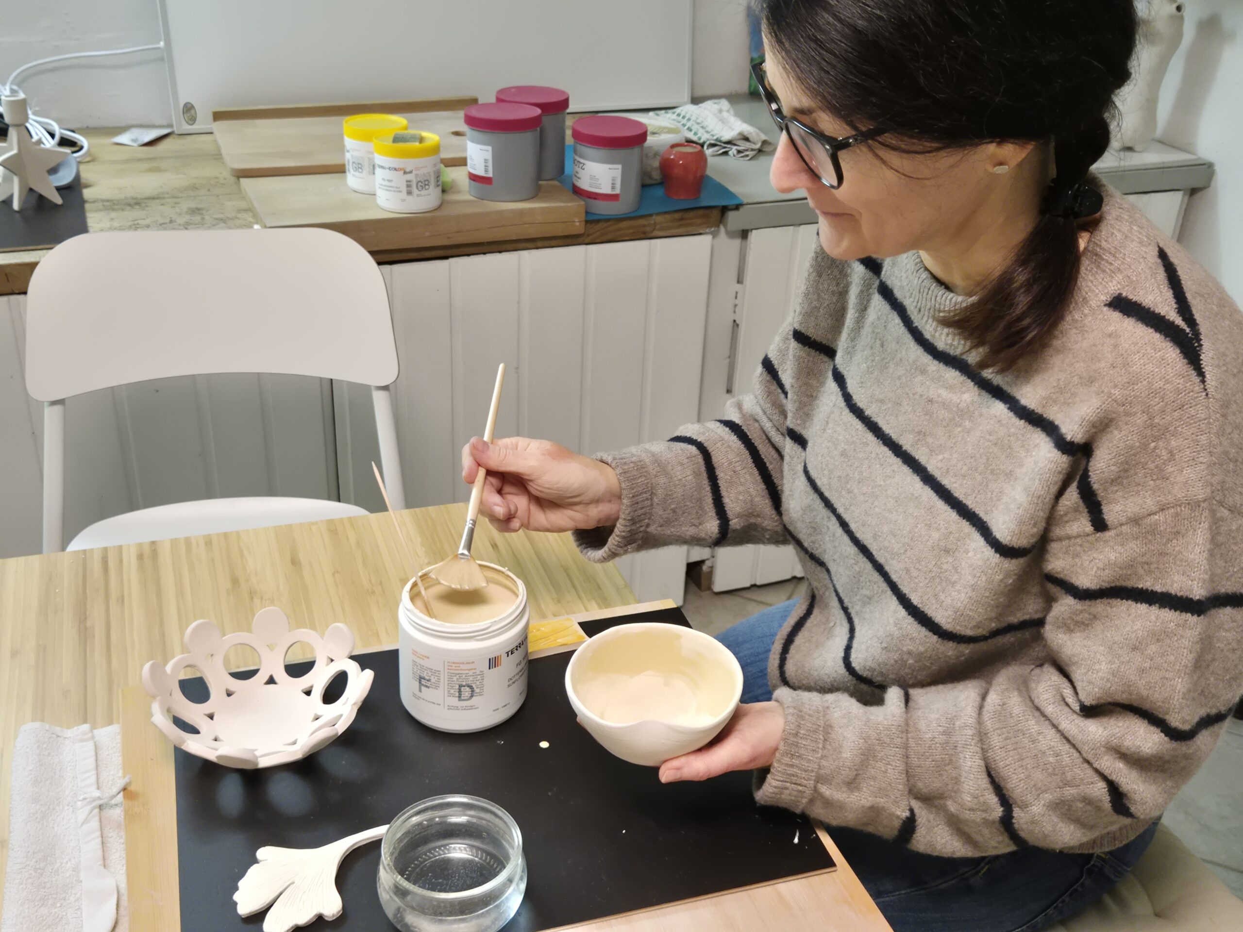 Eine Frau mit Brille und gestreiftem Pullover sitzt an einem Tisch und bemalt eine Keramikschale mit einem Pinsel. Auf dem Holztisch in einem gut beleuchteten Raum stehen Behälter mit Farbe, eine dekorative Keramikfigur und Kunstzubehör.