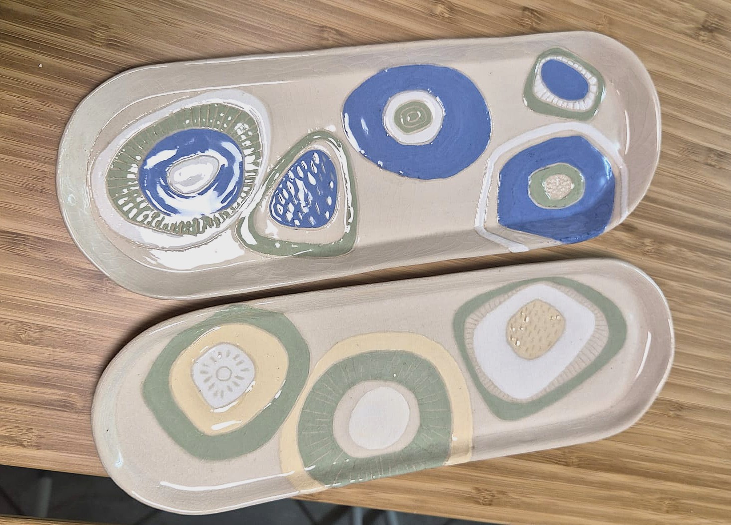 Zwei ovale Keramiktabletts mit abstrakten Kreismustern in Blau, Grün, Beige und Weiß auf einer Holzoberfläche. Die Muster haben ein handgemaltes, organisches Aussehen.