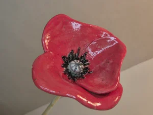Eine Nahaufnahme einer leuchtend roten Mohnblume aus Keramik mit schwarzen Details in der Mitte, die vor einem schlichten, neutralen Hintergrund steht. Die Mohnblume ist auf einem Stab montiert, der inmitten eines Blumenbeetes gesteckt wird und dort mit Wasser befüllt als Bienentränke fungiert.