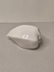 Eine weiße, glatte, abstrakte Keramikskulptur mit einer abgerundeten Form und einem kleinen Vorsprung steht auf einer hellgrauen Oberfläche vor einer strukturierten hellgrauen Wand.