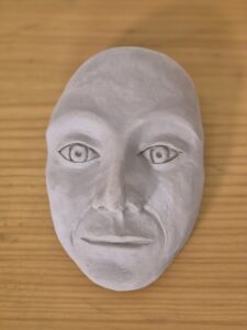 getöpfertes Gesicht, das aussieht wie eine Maske, luftgetrockneter Ton, schmale Lippen, hochgezogene Augenbrauen, dünne Nase