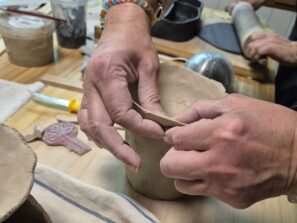 Eine Person formt mit der Hand einen Tontopf auf einem Holztisch, umgeben von Töpferwerkzeugen. Die Szene erinnert an eine praktische Töpfer- oder Keramikwerkstatt.