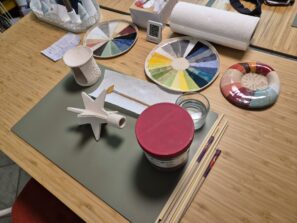 Ein Tischset zum Bemalen von Keramik mit einem weißen Keramikobjekt, Farbkarten, einem Wasserbecher, Pinseln, einem Bleistift, einem Behälter mit rotem Deckel, Papiertüchern und einer handschriftlichen Notiz auf einer Bambusfläche.