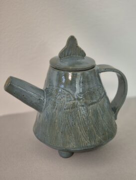 Eine handgefertigte, blaugraue Teekanne aus Keramik mit einem strukturierten, gewellten Design. Der Deckel hat einen einzigartigen, dreieckigen Henkel, und der Körper hat einen runden Sockel mit drei kleinen Füßen. Die Teekanne steht auf einer hellen Unterlage.