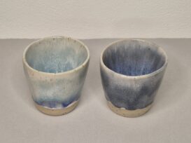 Zwei handgefertigte Keramiktassen mit matter Oberfläche, hellbeigem Grundton und blauer Verlaufsglasur; die linke Tasse ist heller, während die rechte Tasse einen tieferen Blauton aufweist. Beide stehen auf einer hellen Fläche vor einem neutralen Hintergrund.2 Tassen mit gesprenkeltem Ton unten ein feiner Streifen unglasiert oberhalb ist ein Farbverlauf von weiß - mint - blau bei der einen Tasse und bei der anderen von weiß - bis dunkelgrau. Die Tassen haben keinen Henkel.