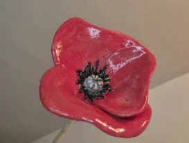Eine Nahaufnahme einer leuchtend roten Mohnblume aus Keramik mit schwarzen Details in der Mitte, die vor einem schlichten, neutralen Hintergrund steht. Die Mohnblume ist auf einem Stab montiert, der inmitten eines Blumenbeetes gesteckt wird und dort mit Wasser befüllt als Bienentränke fungiert.
