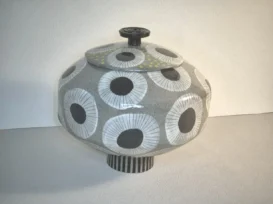 Rundes Keramikgefäß mit Deckel, verziert mit einem Muster aus großen, schwarz-weißen, kreisförmigen Blumenformen und einigen gelben Punkten, das auf einem kurzen, gestreiften Sockel steht.