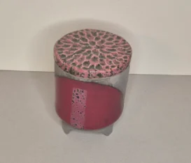 Ein zylindrisches Keramikgefäß mit Deckel. Der Deckel hat ein strukturiertes, strahlenförmiges Muster in Rosa und schwarz gefärbt, während der Körper aus schwarzem Ton gemacht ist und auf einem mit Bordeau gefärbtem Halbkreis eine 1,5cm breite Linie mit schwarzen Sprenkeln ist. Das Gefäß steht auf drei kleinen Füßen.