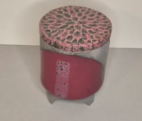 Ein zylindrisches Keramikgefäß mit Deckel. Der Deckel hat ein strukturiertes, strahlenförmiges Muster in Rosa und schwarz gefärbt, während der Körper aus schwarzem Ton gemacht ist und auf einem mit Bordeau gefärbtem Halbkreis eine 1,5cm breite Linie mit schwarzen Sprenkeln ist. Das Gefäß steht auf drei kleinen Füßen.