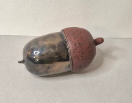 Keramikskulptur einer Eichel mit einem glänzenden, marmorierten braunen und grauen Körper und einem matten, strukturierten rötlich-braunen Hut, der auf einer hellgrauen Oberfläche ruht.