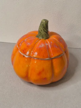 Ein keramischer Behälter in Form und Farbe eines Kürbisses mit orangefarbenem Körper und grünem Stiel, der auf einer hellen Fläche vor einer hellen Wand steht.