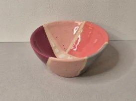 Eine kleine Keramikschale mit glänzender Oberfläche, die geometrische Abschnitte in Rosa, Rot und Grau sowie weiße Akzentlinien auf einem hellen, strukturierten Hintergrund aufweist.
