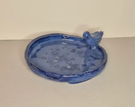 Eine runde, blaue Keramikschale mit einem kleinen, skulptierten Vogel auf dem Rand. Die Schale hat einen glatten, leicht unebenen Rand und verstreute kreisförmige Muster auf dem Boden.