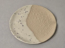 Ein runder Keramikteller mit einem diagonal geteilten Muster: eine Hälfte ist glatt und cremefarben mit dunklen Sprenkeln, die andere Hälfte hat ein strukturiertes, beigefarbenes Muster, das an Spitze oder Schuppen erinnert. Der Teller ruht auf einer hellgrauen Fläche.