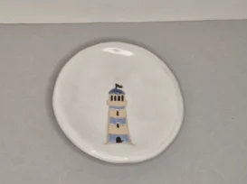 Ein runder weißer Keramikteller zeigt eine einfache Illustration eines beigen und blauen Leuchtturms mit kleinen Fenstern, zentriert auf einem schlichten grauen Hintergrund.