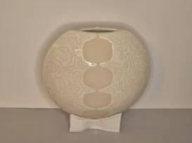 Runde, cremefarbene Keramikvase mit schmaler Öffnung, verziert mit erhabenen, gewellten Linien und einer vertikalen Säule aus ovalen Formen in der Mitte, die auf einem kleinen, skulptierten Sockel steht.