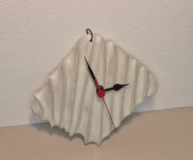Eine Wanduhr mit gewelltem, unregelmäßigem, weißem Keramikzifferblatt, schwarzem Stunden- und Minutenzeiger und rotem Sekundenzeiger, die vor einer hellen Wand hängt.